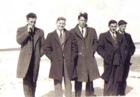 Five men in suits and overcoats stand together along a bright, wintry shoreline.