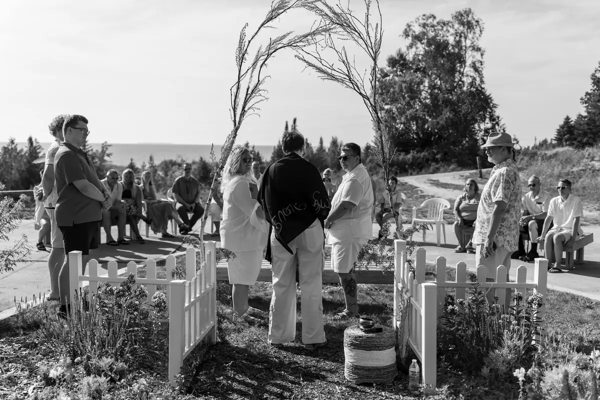 Black-and-white outdoor ceremony with seated attendees beside a small flower garden and body of water.