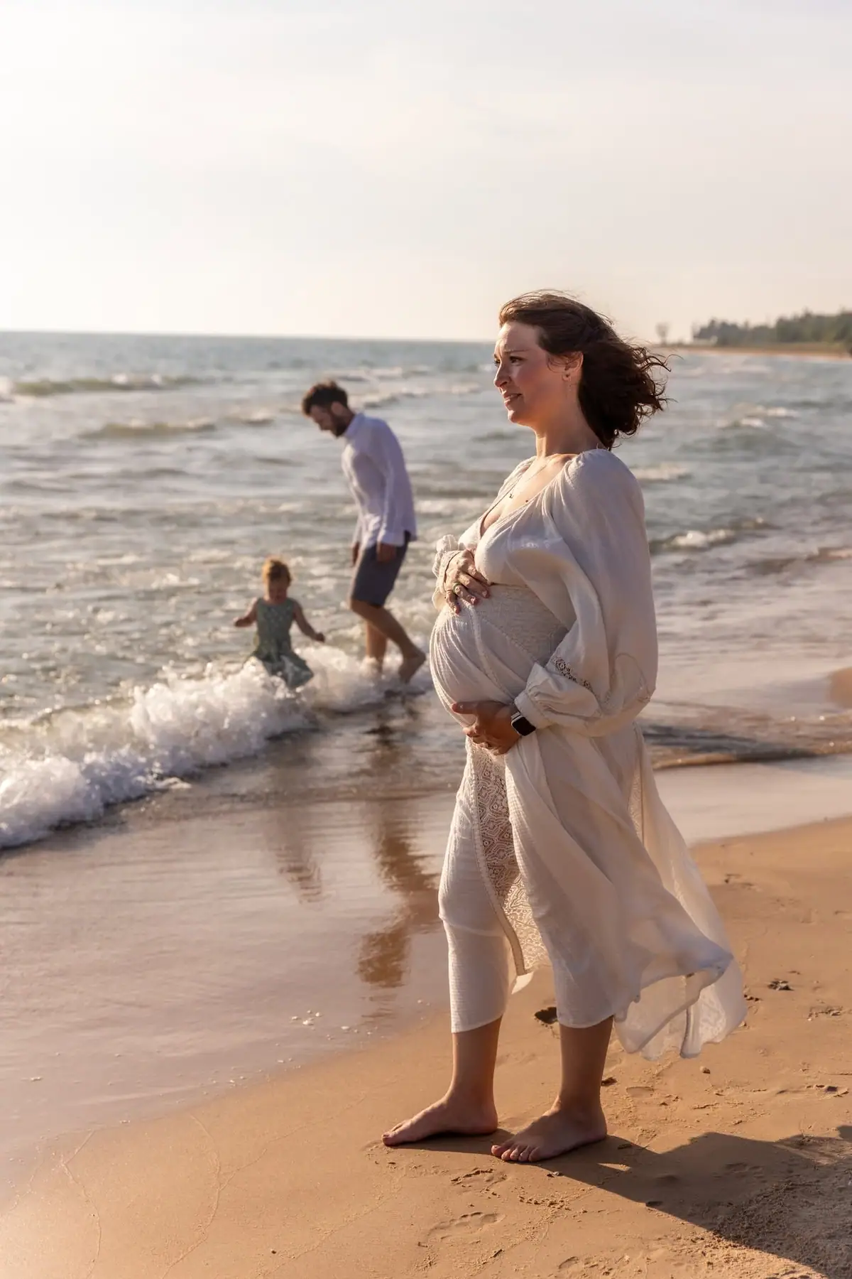 A pregnant woman in a flowing white outfit stands barefoot on a sandy beach while two people play in the waves.