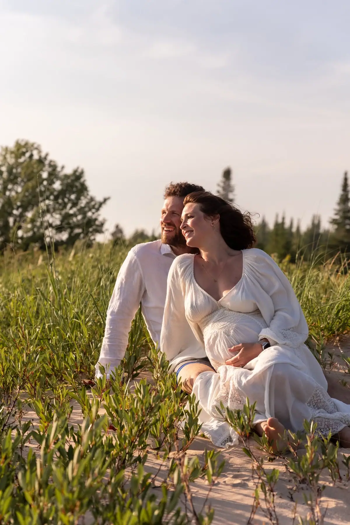 A couple in white clothing sits among grasses and low shrubs on sandy ground beneath a pale sky.