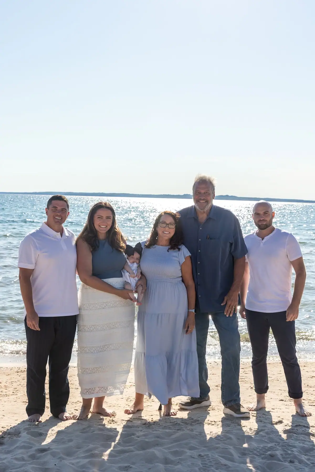 Five adults and an infant pose together on a sandy beach beside sparkling water.
