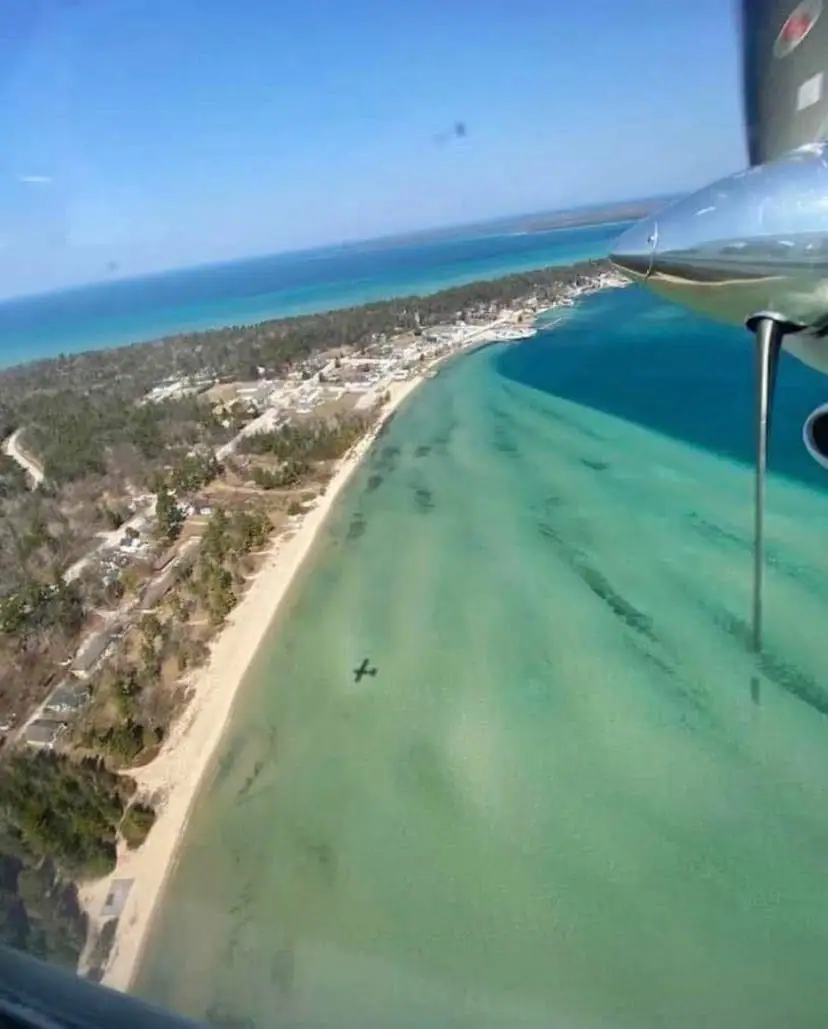 Aerial view of a sandy shoreline, turquoise water, wooded settlement, and a propeller aircraft engine.