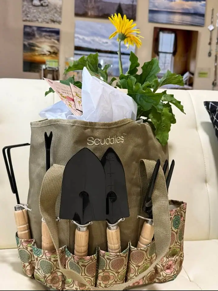 A canvas gardening tote holds hand tools and a leafy plant with a bright yellow flower.