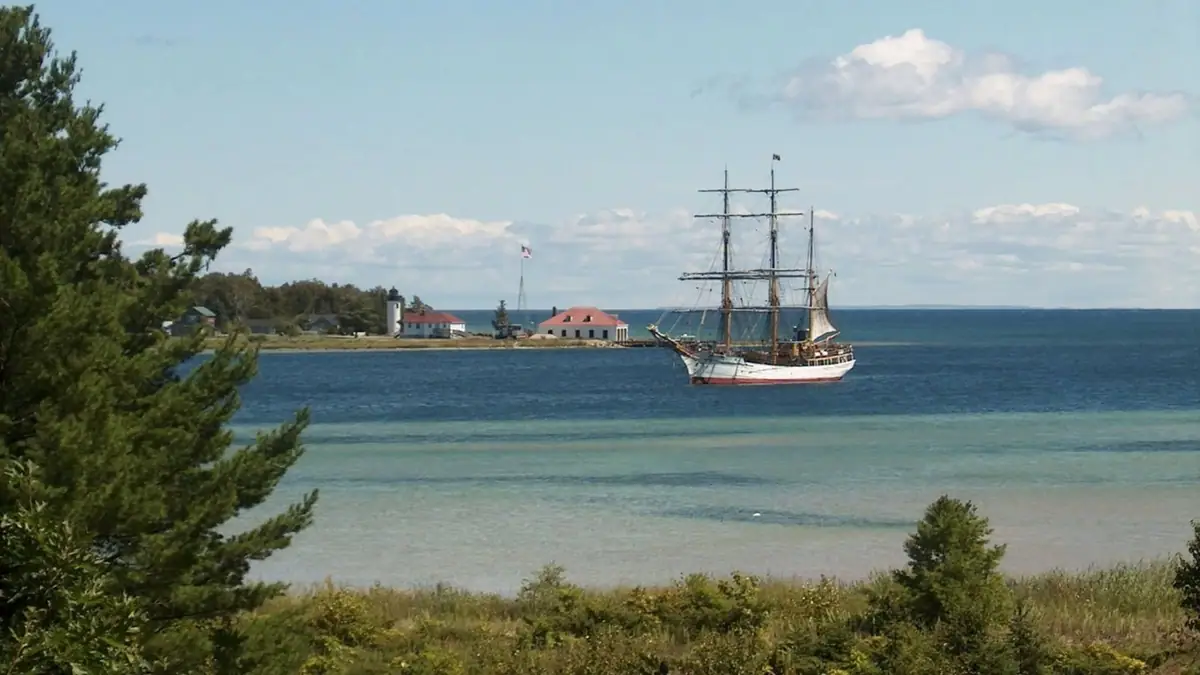 Three-masted sailing ship anchored offshore near a lighthouse and shoreline buildings.