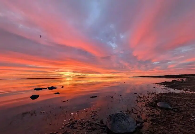 A vivid sunset spreads across calm water beside a rocky shoreline.