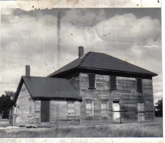 Black-and-white photograph of a weathered two-story wooden house with a smaller attached wing.