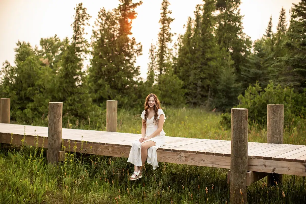 A smiling woman in a white dress sits on a wooden boardwalk amid tall grasses and trees.