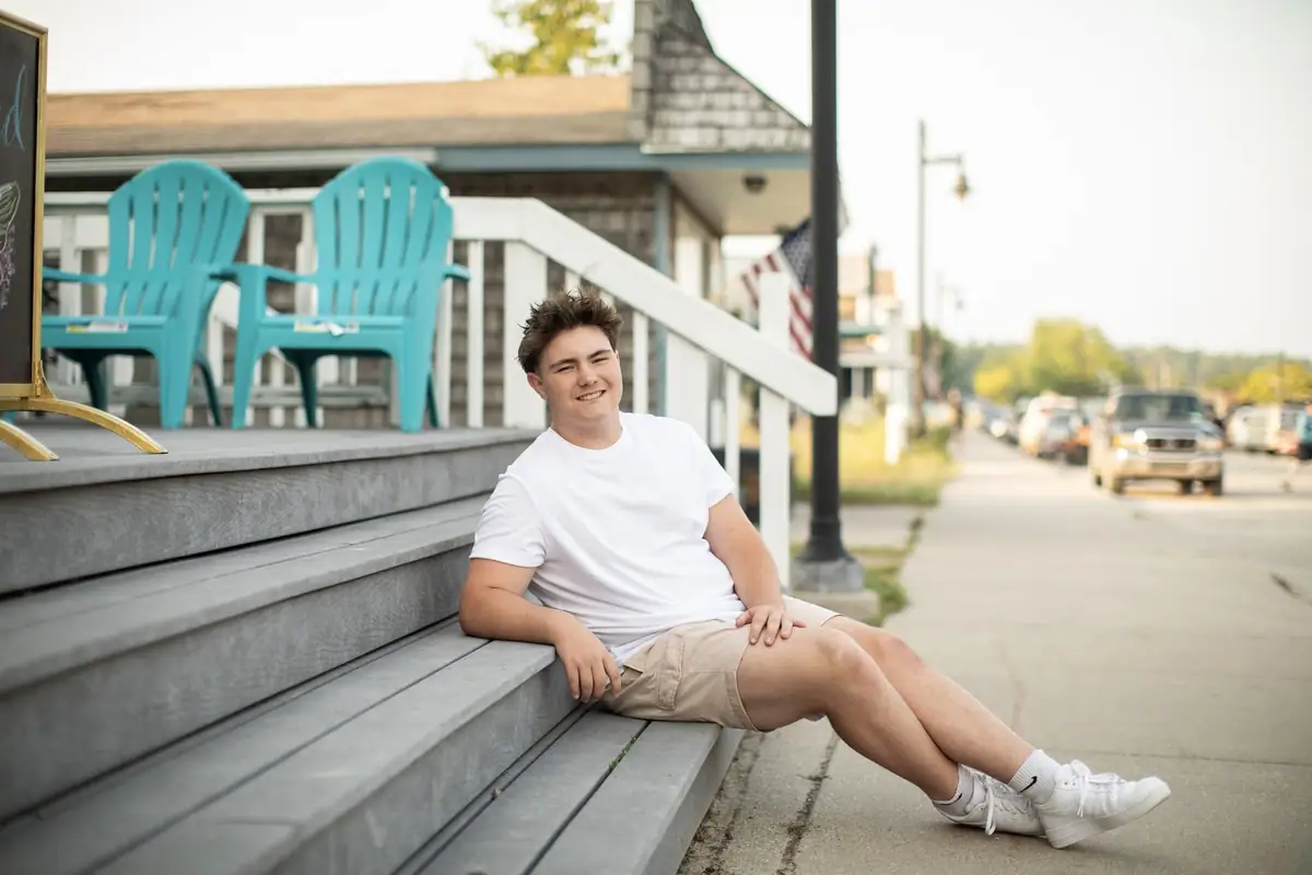 A young man sits on broad wooden steps beside blue chairs and a sidewalk, with a flag and parked vehicles behind him.