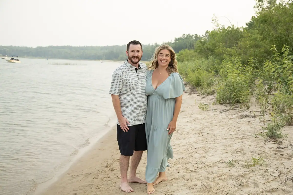 A smiling couple poses barefoot on a sandy shore beside calm water and a wooded opposite shoreline.