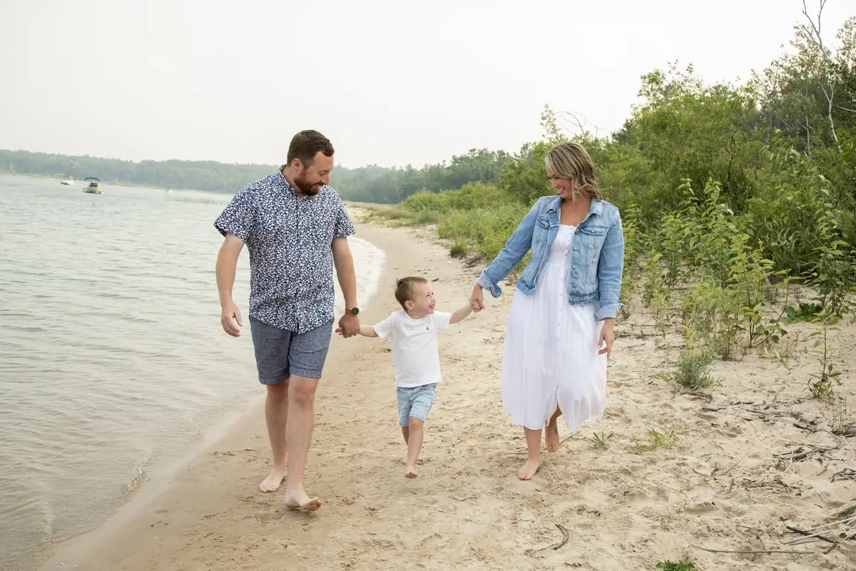 Three people walk barefoot along a sandy shoreline beside calm water and wooded vegetation.