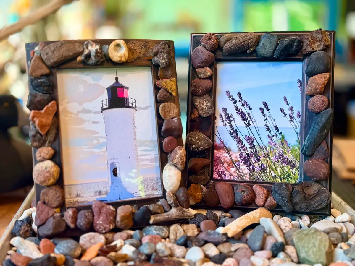 Two stone-decorated frames display a lighthouse image and purple flowers above a bed of colorful stones and shells.