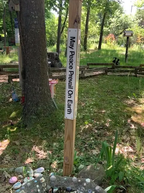 A wooden post with a peace message stands among trees, benches, birdhouses, and decorated stones.