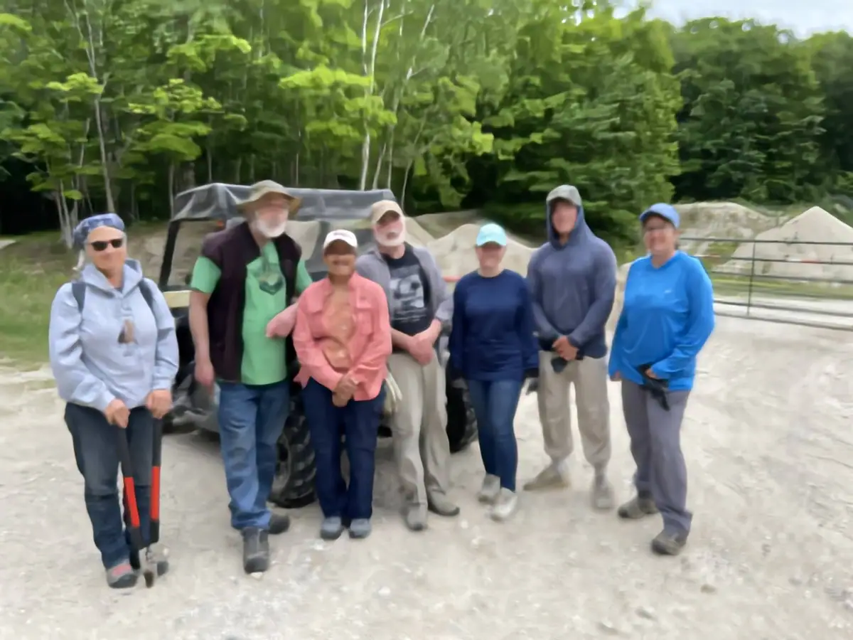 Seven people pose beside a dark utility vehicle on a gravel clearing surrounded by forest.