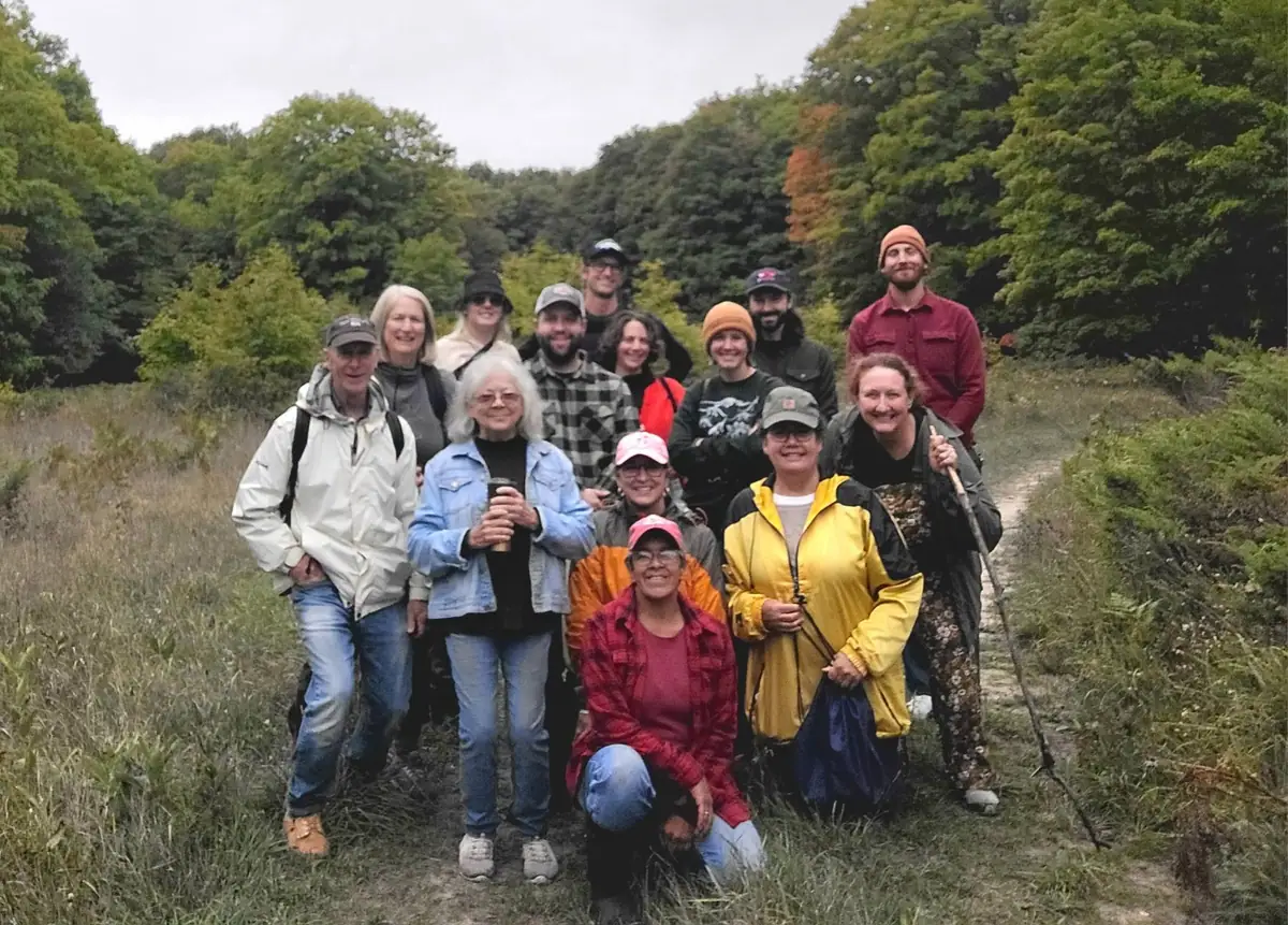 A group of people poses on a wooded trail surrounded by dense green trees and patches of autumn color.