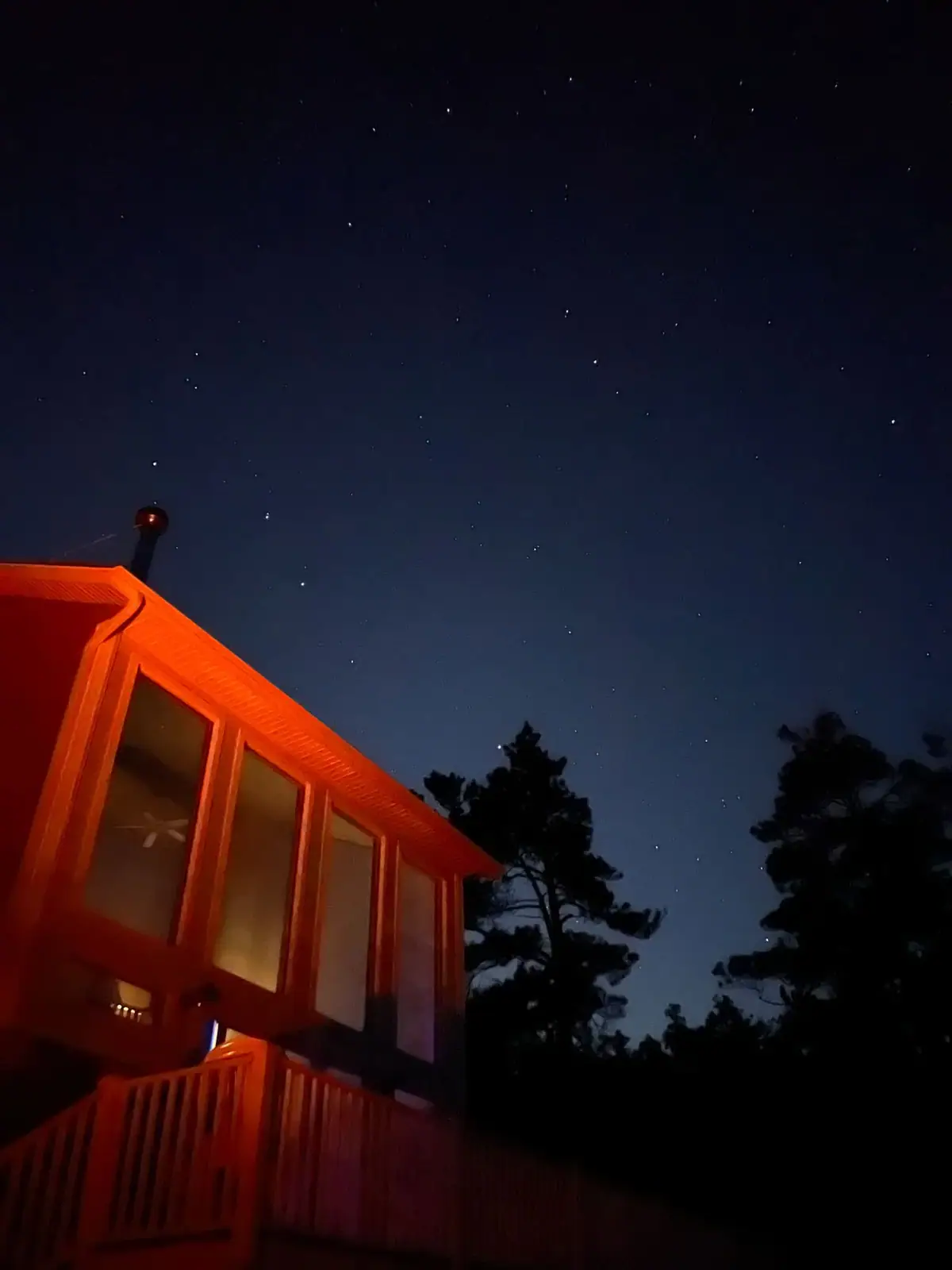A warmly lit house and deck stand beneath a star-filled night sky and silhouetted trees.