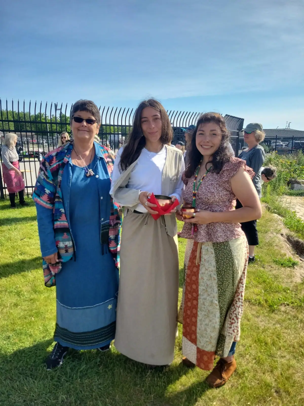 Three women pose outdoors in colorful clothing while holding small vessels near a black metal fence.