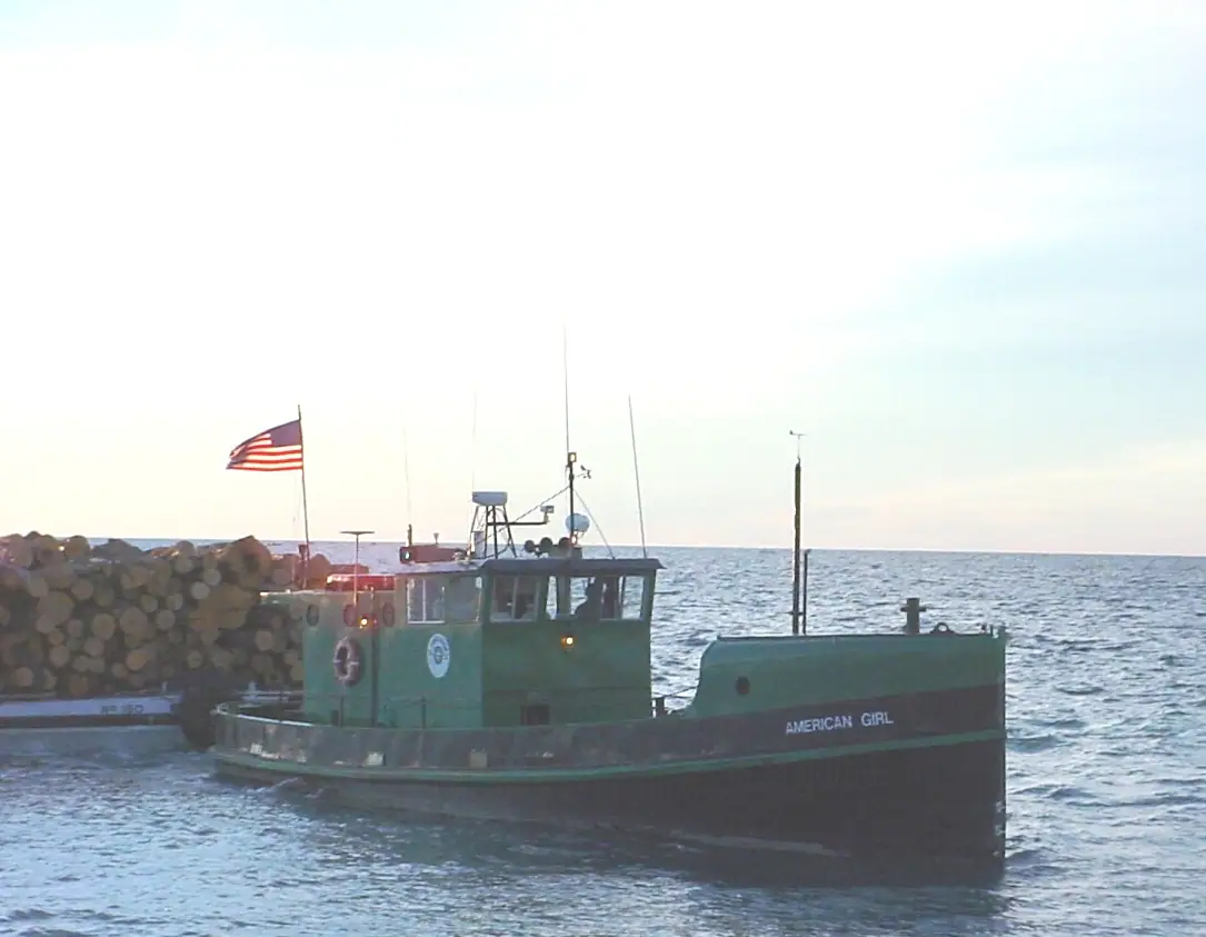 A green tugboat towing a barge loaded with logs across open water.
