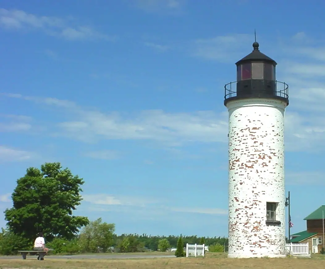 A weathered white lighthouse rises beside a green-roofed building under a blue sky.