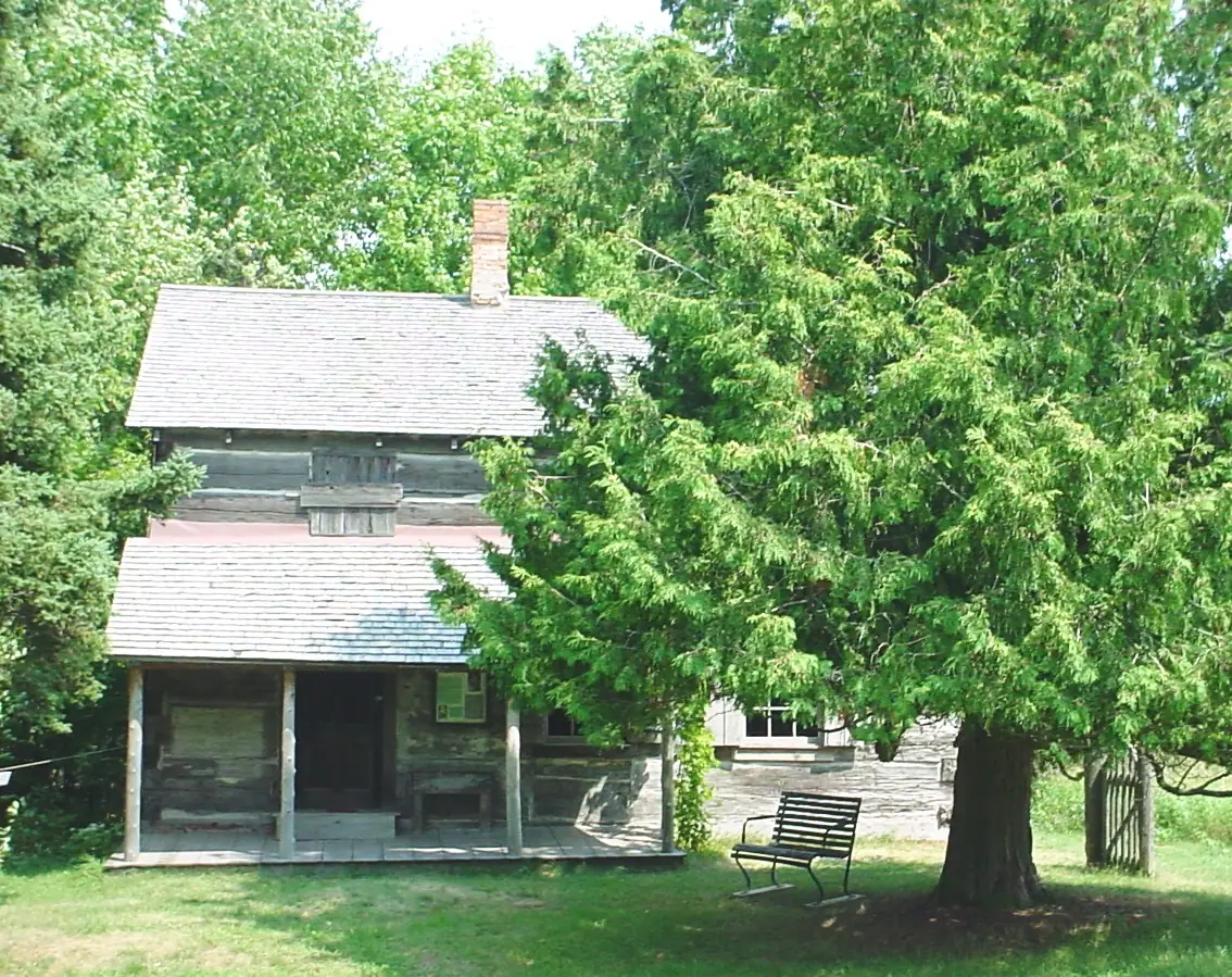 A log house with a covered porch stands among trees beside a bench and a large evergreen.