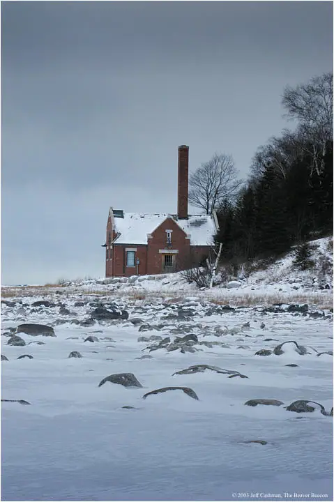 A red brick house with a tall chimney stands beside a snow-covered, rocky shoreline.