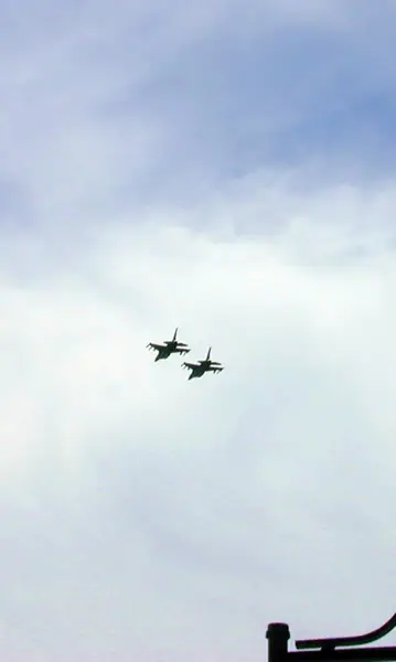 Two fighter jets fly in formation beneath a cloudy sky above a dark rooftop silhouette.