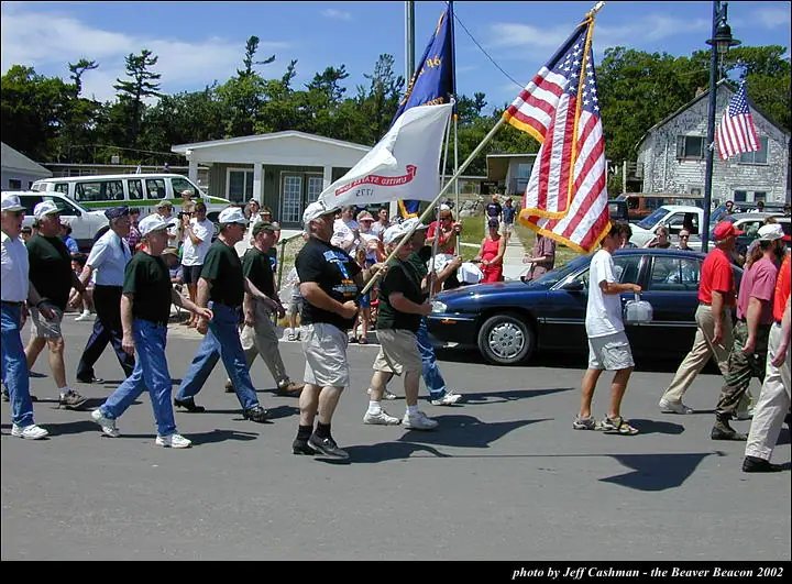 Marchers carrying American and ceremonial flags proceed along a paved street as spectators watch from beside parked cars.