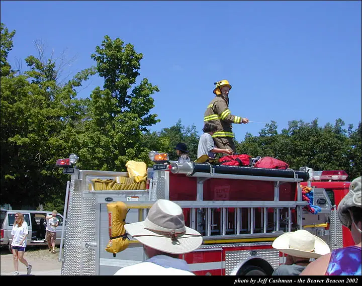 A firefighter stands atop a red fire truck as people gather nearby.
