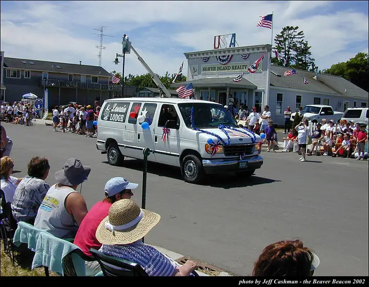 A decorated white van passes seated spectators during a parade near a flag-decorated building.