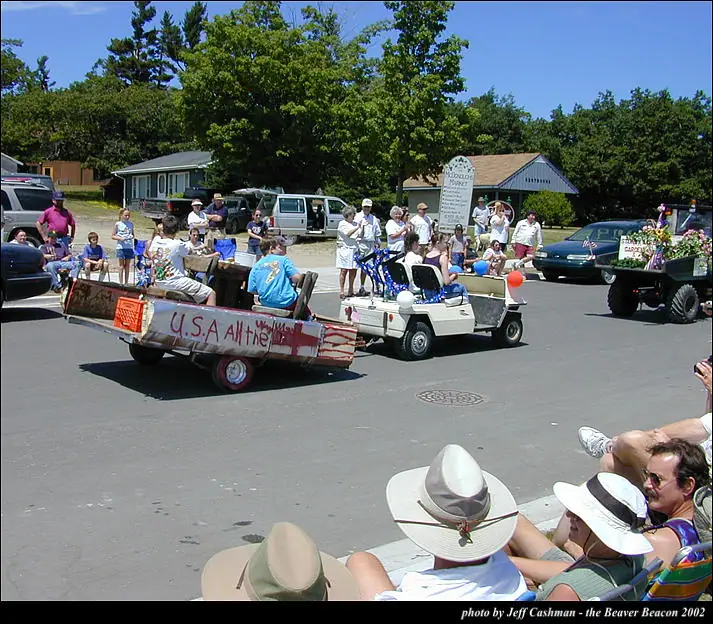 A decorated trailer and golf cart pass spectators during a parade on a sunny paved street.