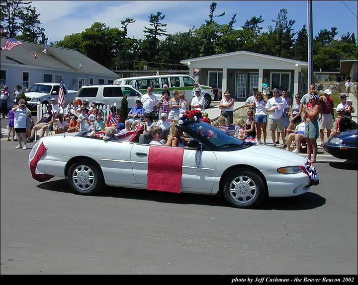 A decorated white convertible passes a crowd during a parade, with flags and a pink banner on its side.