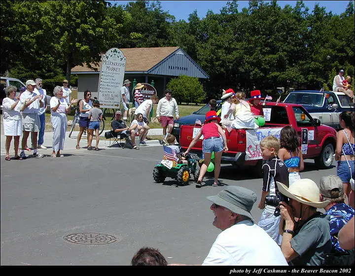 A parade scene with spectators, a decorated red pickup truck, and a child riding a green ATV with an American flag.