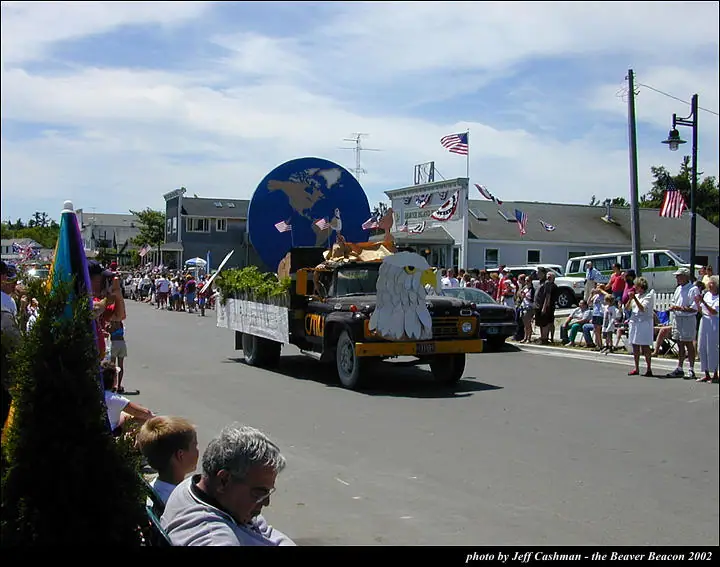 A decorated military-style truck leads a flag-filled parade past spectators on a sunny street.