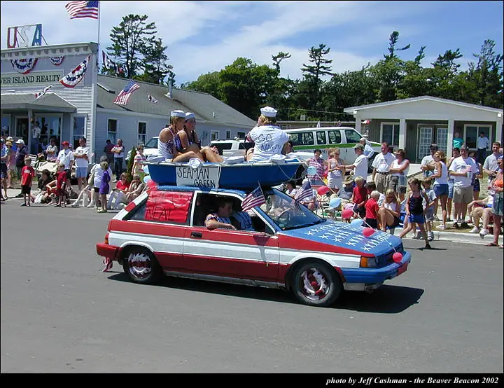 A patriotic parade float carries a small boat on a decorated automobile as spectators watch from the street.
