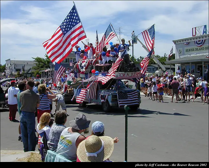A patriotic parade float covered in American flags moves past spectators gathered along a street.