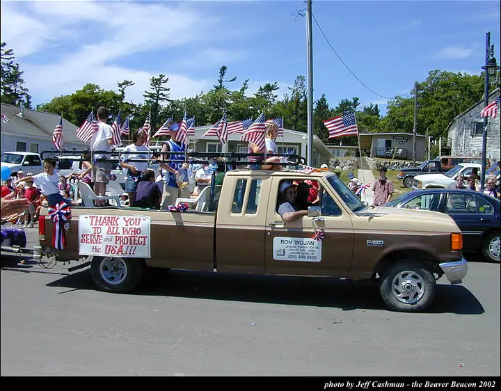 A brown pickup truck carries people and American flags in a parade, with a sign thanking those who serve and protect.