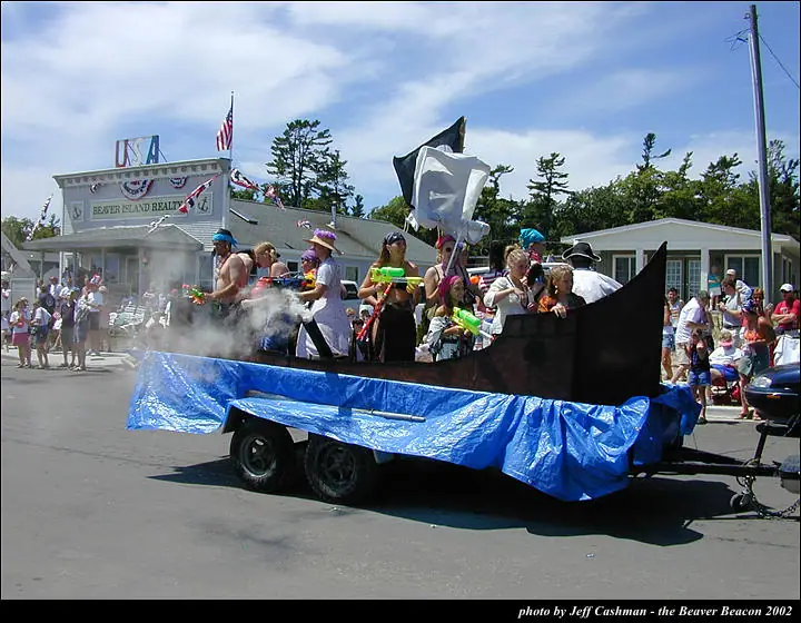 A boat-shaped parade float with people holding water pistols passes spectators on a sunny street.