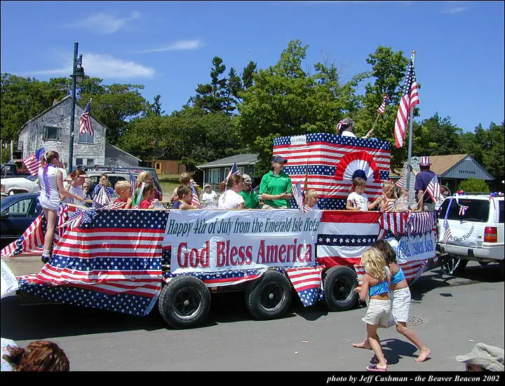 Patriotic parade float covered in American flags and bunting, carrying adults and children along a residential street.