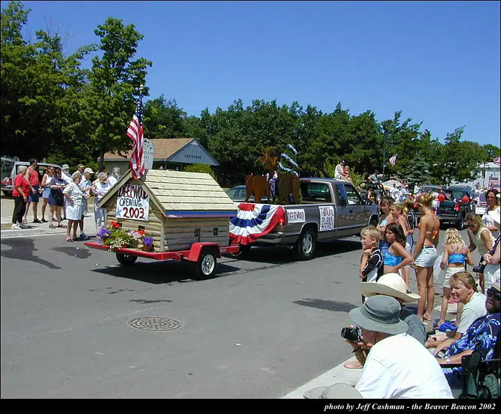 A decorated pickup truck pulls a small cabin-like float past spectators during a sunny street parade.