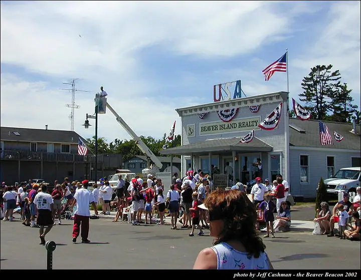 Crowd gathered outside a decorated realty office during a patriotic parade, with a boom lift and flags visible.