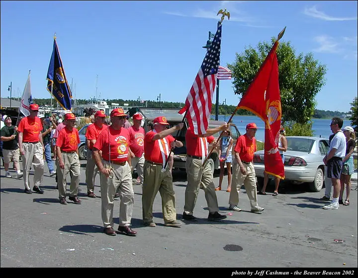 Men in matching red shirts and khaki trousers march with flags along a waterfront road.