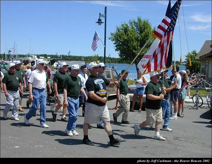 Adults march along a waterfront street carrying American flags and a large patriotic banner.