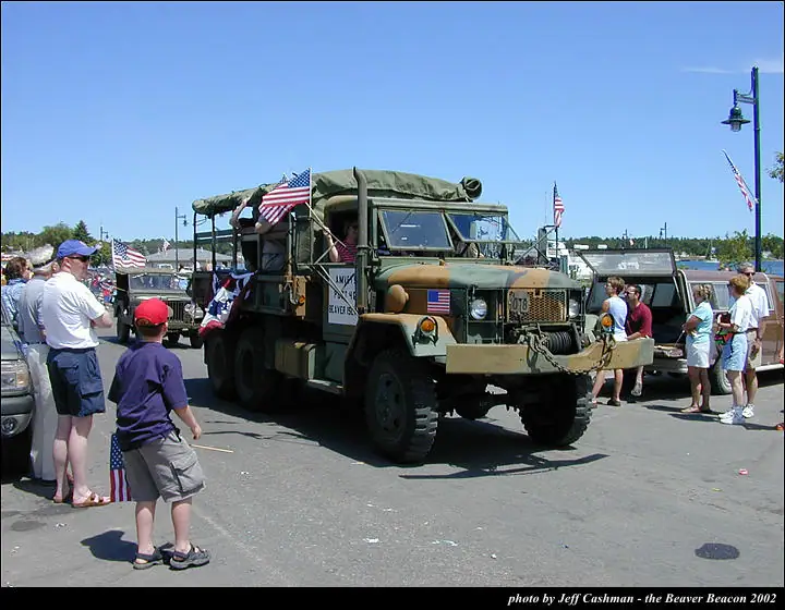 A military-style truck decorated with American flags is surrounded by spectators on a sunny street.