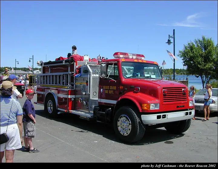 A red fire engine is surrounded by spectators beside a waterfront, with people seated on top of the vehicle.