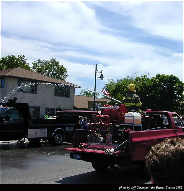 A firefighter rides on a red fire truck during a street parade with another truck and an American flag nearby.