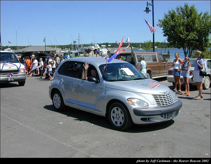 A decorated silver car passes spectators in a waterfront parade.