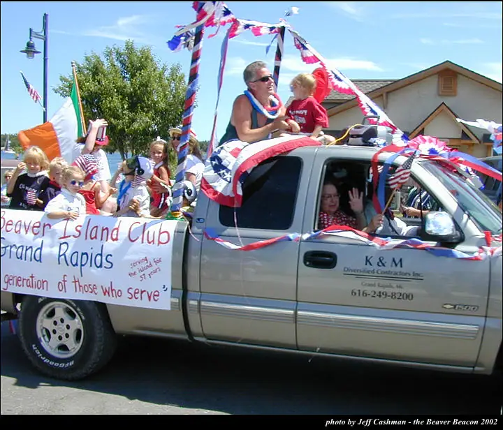Decorated pickup truck carrying adults and children in a parade, with patriotic streamers and a large club banner.