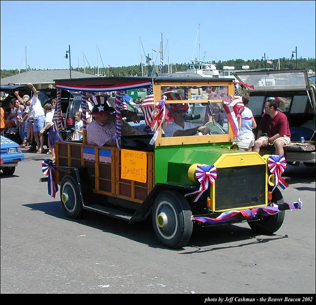 A brightly decorated vintage automobile with passengers and patriotic bunting moves past sailboats and spectators.