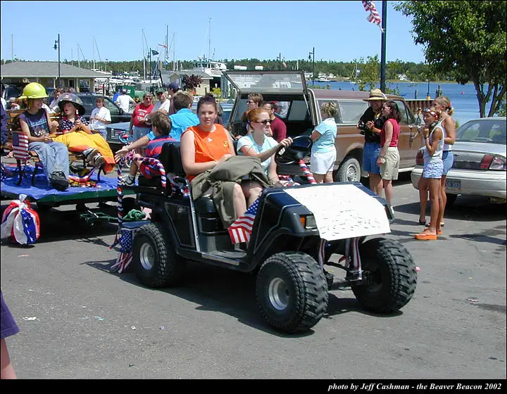 People ride in a flag-decorated utility cart and trailer beside a marina during a parade.
