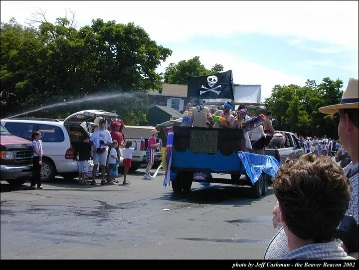 A decorated pickup truck carries costumed people beneath a pirate flag as water sprays across a parade crowd.