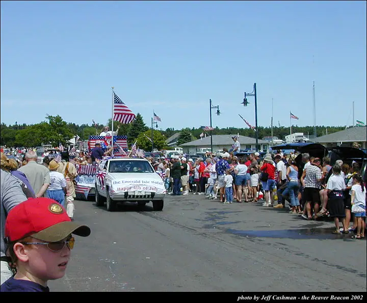 A decorated white SUV leads a flag-filled parade past crowds lining a paved street.